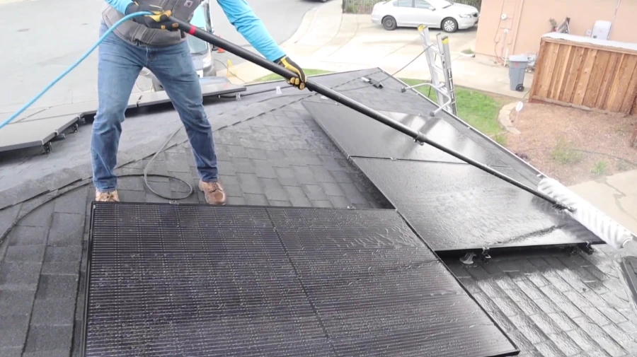 Technician cleaning black solar panels on a two-story shingle roof with a water-fed pole